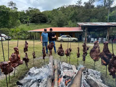 Dos hombres posan junto a un asado sobre brasas con la casa de la finca al fondo
